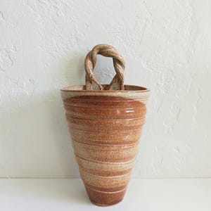May include: A cone-shaped ceramic wall pocket with a speckled brown and tan glaze. The vessel features horizontal ridges along its body and a single, thick, twisted handle at the top center for hanging, showcasing a rustic, earthy aesthetic.