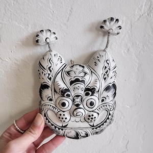 May include: A white and black decorative mask with a detailed, symmetrical design. The mask features a stylized face with large eyes, a prominent nose, and decorative elements. Two floral-like accents extend from the top. The mask is held against a white textured wall.