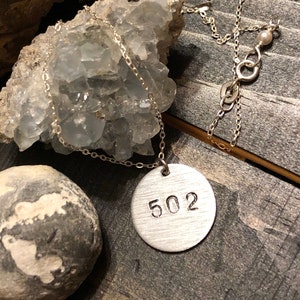 May include: A silver necklace with a circular pendant stamped with the number "502". The necklace chain is delicate, and a small pearl is near the clasp. The pendant is displayed on a rough, textured surface.