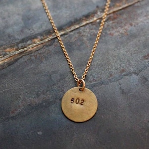Peut inclure: Un délicat collier doré avec un pendentif rond. Le pendentif porte le numéro "502" estampé. La chaîne du collier est fine et le fermoir est visible. Le fond est une surface texturée gris foncé.