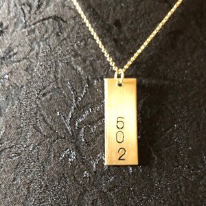 Peut inclure: Un collier doré avec un pendentif rectangulaire. Le pendentif porte le numéro "502" gravé. La chaîne du collier est de couleur dorée délicate, sur un fond de tissu sombre et texturé.