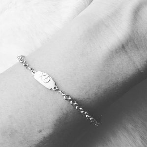 May include: A silver bracelet with an oval charm engraved with "XO" is displayed. The bracelet features a beaded chain and is worn on a wrist. The image is in black and white.