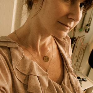 Może przedstawiać: Warstwowy złoty naszyjnik z małą zawieszką i okrągłą zawieszką z numerem "602" jest noszony na jasnor&oacute;żowej bluzce z falbanami. Bluzka ma miękki, płynny design.