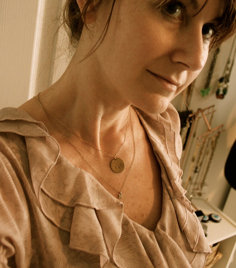 Może przedstawiać: Warstwowy złoty naszyjnik z małą zawieszką i okrągłą zawieszką z numerem "602" jest noszony na jasnor&oacute;żowej bluzce z falbanami. Bluzka ma miękki, płynny design.