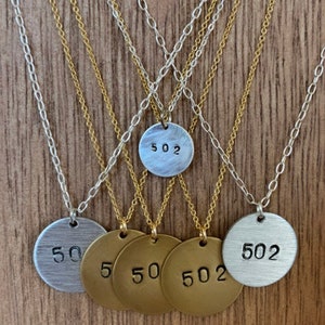 Może przedstawiać: Kolekcja naszyjnik&oacute;w z okrągłymi wisiorkami. Wisiorki są srebrne i złote, z wytłoczonymi numerami "502" i "50". Naszyjniki mają delikatne łańcuszki, niekt&oacute;re srebrne, a niekt&oacute;re złote, na tle drewnianym.