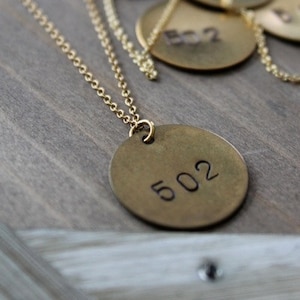 Peut inclure: Un collier doré avec un pendentif circulaire gravé du numéro "502". Le pendentif est suspendu à une chaîne délicate. D'autres pendentifs similaires avec des numéros sont visibles en arrière-plan.