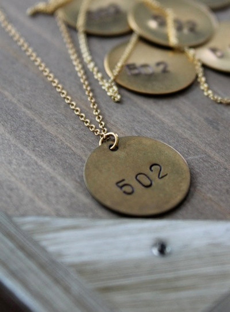 Może przedstawiać: Złoty naszyjnik z okrągłą zawieszką z wygrawerowanym numerem "502". Zawieszka wisi na delikatnym łańcuszku. W tle widoczne są inne podobne zawieszki z numerami.