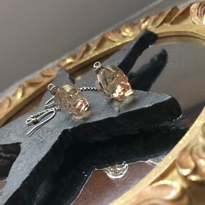 May include: A pair of skull-shaped earrings. Each earring features a small, faceted, amber-colored crystal skull suspended from a silver-colored chain and hook. The earrings are displayed on a dark, textured surface.