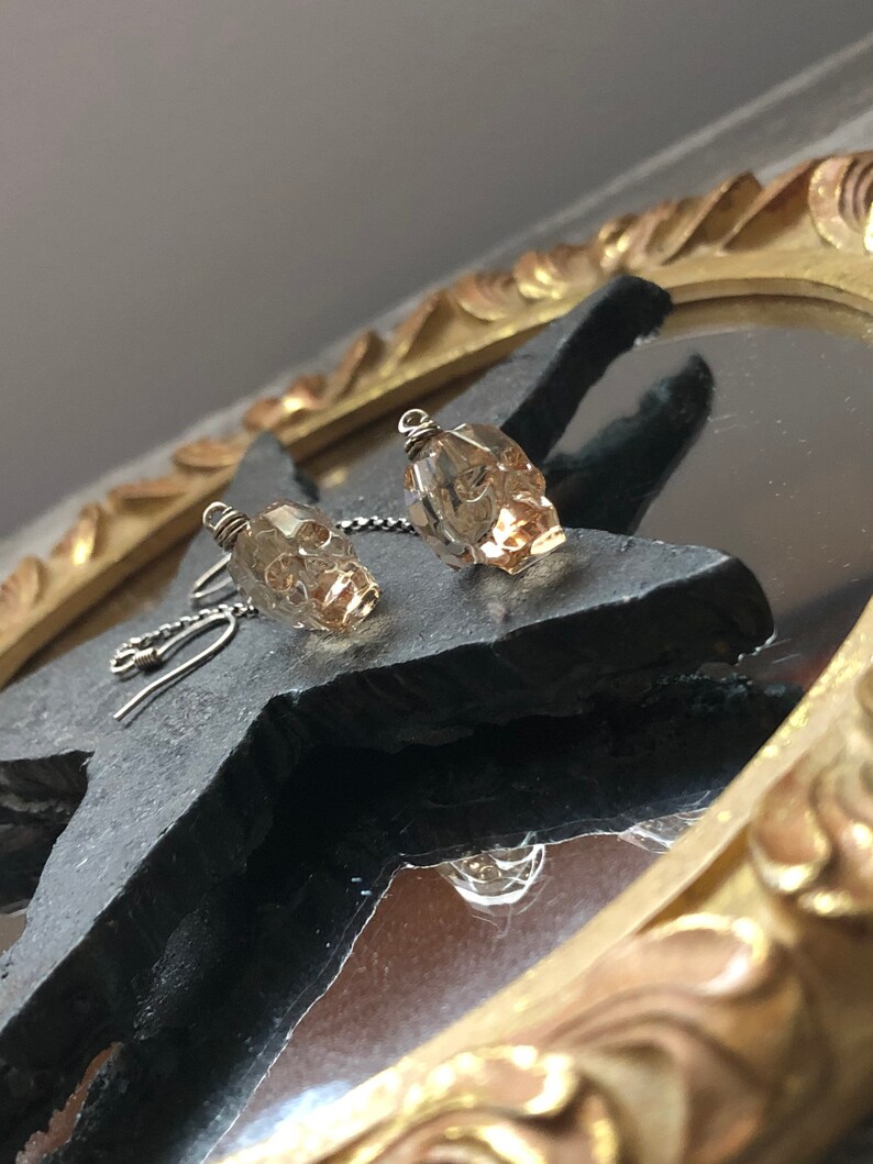 May include: A pair of skull-shaped earrings. Each earring features a small, faceted, amber-colored crystal skull suspended from a silver-colored chain and hook. The earrings are displayed on a dark, textured surface.