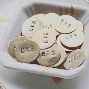 Może przedstawiać: Kolekcja małych, okrągłych, mosiężnych metalowych zawieszek, każda z numerem "513". Zawieszki są ułożone w białej, kwadratowej misce z lekko zakrzywionymi krawędziami. Zawieszki mają matowe wykończenie.