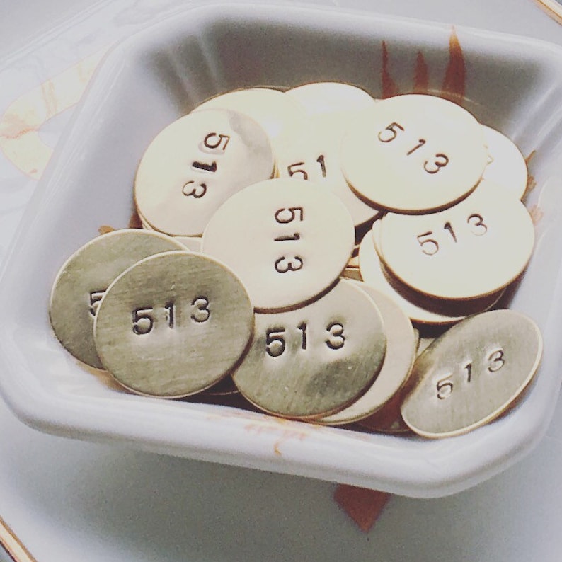 Może przedstawiać: Kolekcja małych, okrągłych, mosiężnych metalowych zawieszek, każda z numerem "513". Zawieszki są ułożone w białej, kwadratowej misce z lekko zakrzywionymi krawędziami. Zawieszki mają matowe wykończenie.