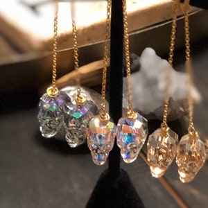 May include: Gold-colored chain earrings with skull-shaped crystal pendants. The crystals have a multifaceted design, reflecting light with iridescent colors. The earrings are displayed against a dark background.
