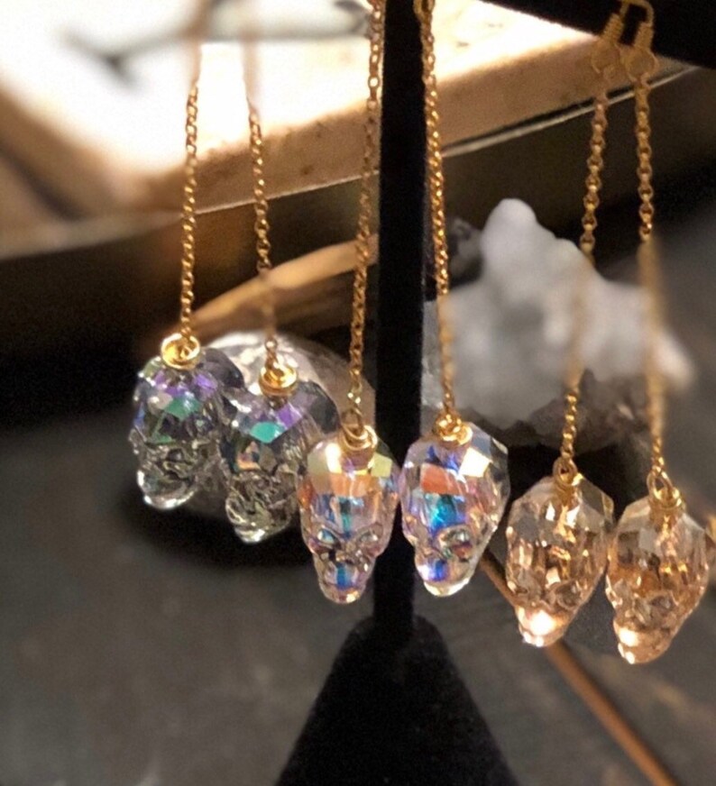 May include: Gold-colored chain earrings with skull-shaped crystal pendants. The crystals have a multifaceted design, reflecting light with iridescent colors. The earrings are displayed against a dark background.