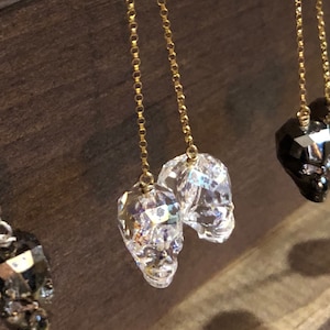 May include: Close-up of several pairs of dangle earrings. The earrings feature faceted skull charms in clear and black, suspended from delicate gold-colored chains. The background is a dark wood surface.