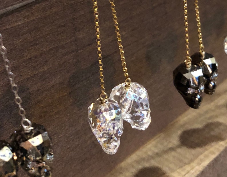 May include: Close-up of several pairs of dangle earrings. The earrings feature faceted skull charms in clear and black, suspended from delicate gold-colored chains. The background is a dark wood surface.