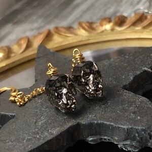 May include: A pair of skull-shaped earrings. The earrings are dark gray with a metallic sheen, suspended from gold-colored hooks and chains. The skulls have detailed features and are wire-wrapped at the top.