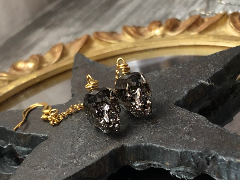 May include: A pair of skull-shaped earrings. The earrings are dark gray with a metallic sheen, suspended from gold-colored hooks and chains. The skulls have detailed features and are wire-wrapped at the top.