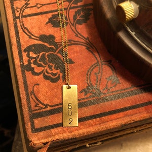May include: A gold-colored rectangular pendant necklace with the number "502" engraved on it. The pendant hangs from a delicate chain. The necklace rests on a vintage book with floral designs.