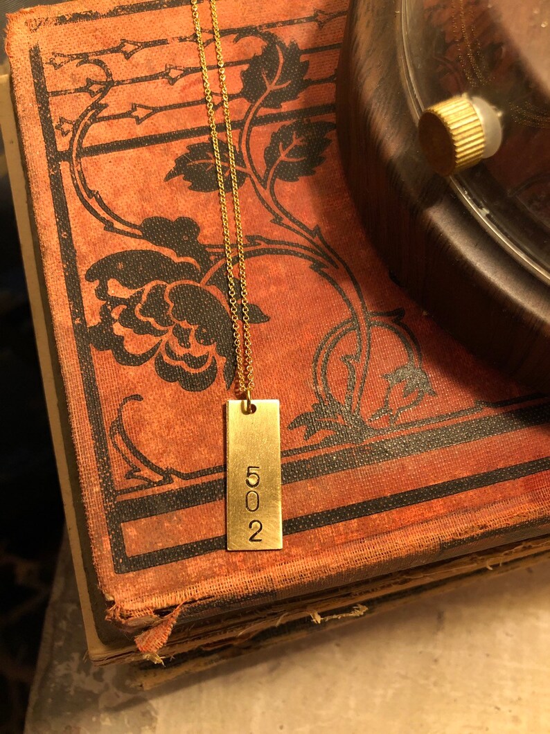 May include: A gold-colored rectangular pendant necklace with the number "502" engraved on it. The pendant hangs from a delicate chain. The necklace rests on a vintage book with floral designs.