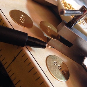 Może przedstawiać: Zbliżenie narzędzi i materiał&oacute;w do tworzenia biżuterii. Czarny długopis znajduje się nad złotą owalną etykietą z numerem "606". Widoczne są inne etykiety z "909", a także linijka i małe złote koraliki.