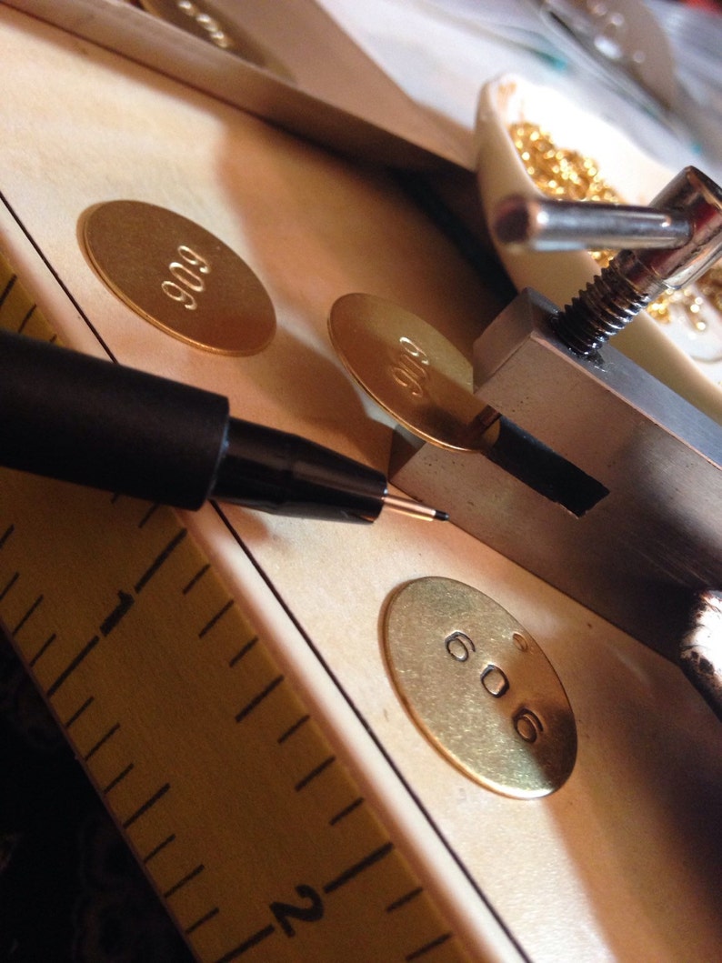 Może przedstawiać: Zbliżenie narzędzi i materiał&oacute;w do tworzenia biżuterii. Czarny długopis znajduje się nad złotą owalną etykietą z numerem "606". Widoczne są inne etykiety z "909", a także linijka i małe złote koraliki.