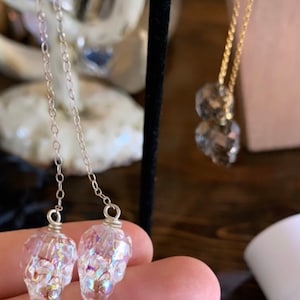 May include: Clear skull-shaped earrings with iridescent glitter suspended inside, hanging from delicate silver chains. A gold necklace with two faceted crystal pendants is also visible. The jewelry is displayed on a dark background.