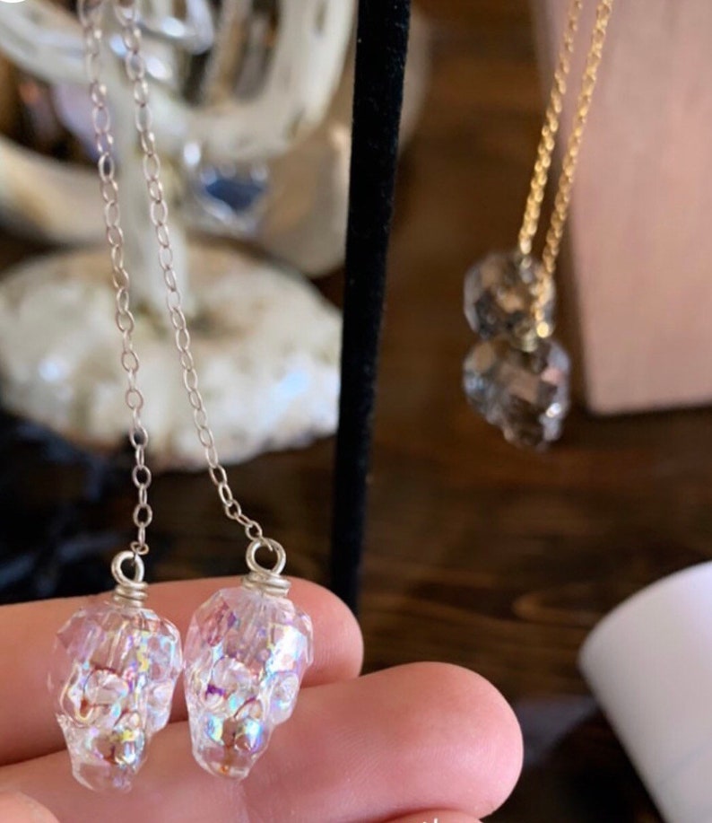 May include: Clear skull-shaped earrings with iridescent glitter suspended inside, hanging from delicate silver chains. A gold necklace with two faceted crystal pendants is also visible. The jewelry is displayed on a dark background.