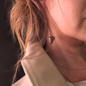 May include: A gold-colored earring with a skull pendant hangs from the ear. The earring is suspended by a delicate chain. The background shows a person's hair and part of a white jacket.