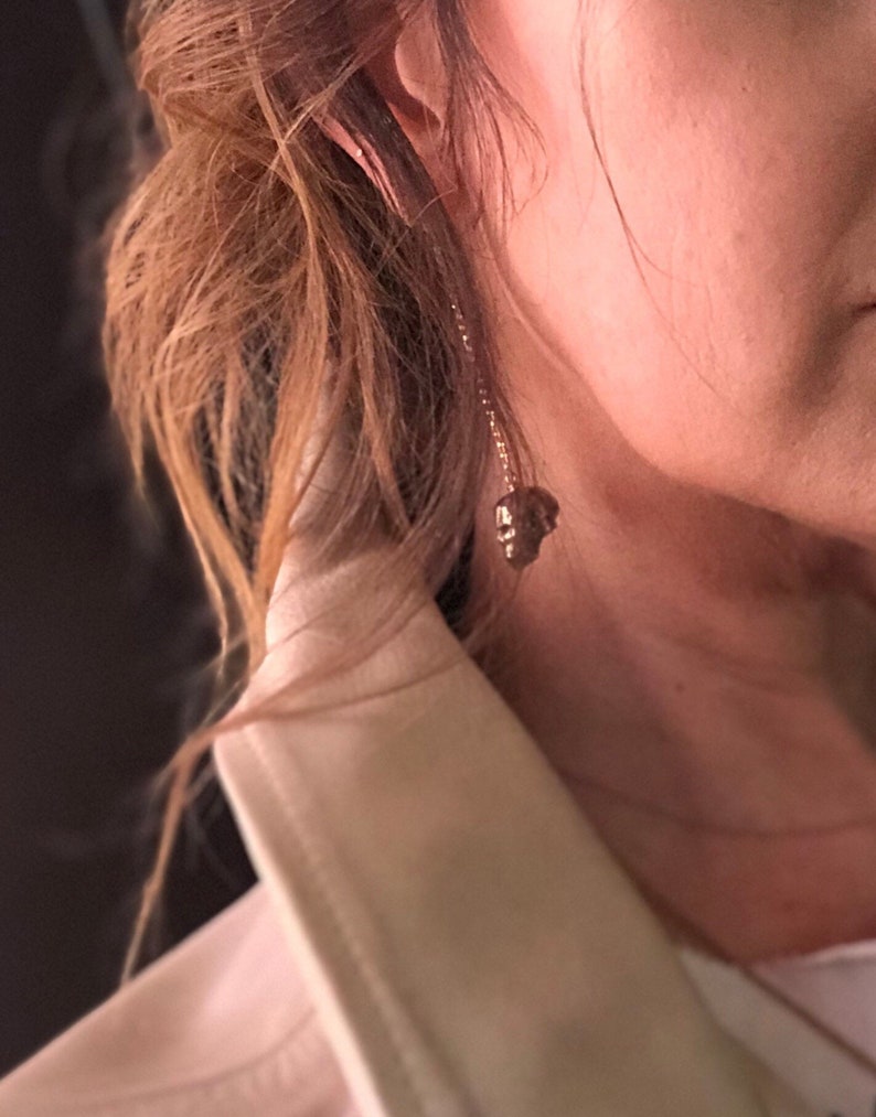 May include: A gold-colored earring with a skull pendant hangs from the ear. The earring is suspended by a delicate chain. The background shows a person's hair and part of a white jacket.