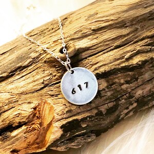 Peut inclure: Un collier argenté avec un pendentif rond portant le numéro "617". Le pendentif est suspendu à une chaîne fine. L'ensemble est présenté sur un fond de bois vieilli.