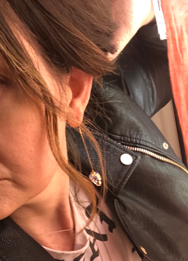 May include: A gold-colored chain earring with a large, iridescent crystal pendant. The crystal reflects multiple colors. The earring is dangling near a person's ear. The person is wearing a black leather jacket.