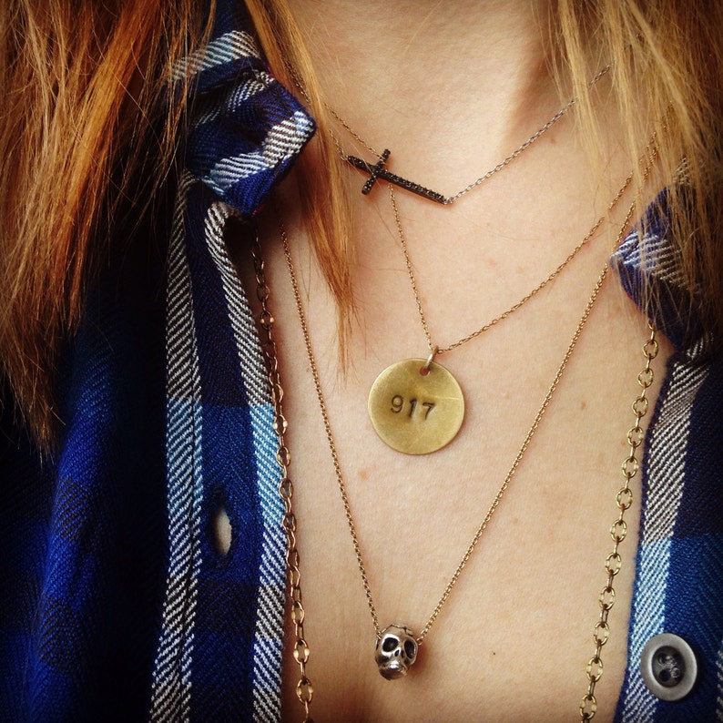 Może przedstawiać: Zestaw naszyjnik&oacute;w warstwowych z czarną zawieszką w kształcie krzyża, złotą zawieszką w kształcie dysku z numerem "917" i zawieszką w kształcie czaszki. Naszyjniki są noszone na niebiesko-białej koszuli w kratę.