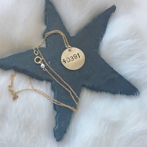 Puede incluir: Un collar dorado con un colgante circular con el número "40391". El colgante cuelga de una delicada cadena dorada. El collar se exhibe sobre un objeto en forma de estrella gris oscuro, sobre un fondo blanco y esponjoso.