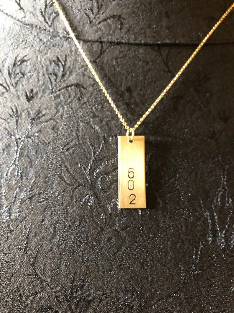 May include: A gold-colored necklace with a rectangular pendant. The pendant has the number "502" engraved on it. The necklace chain is a delicate gold color, and the background is a dark, textured fabric.