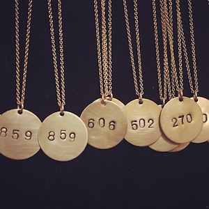 May include: A collection of gold-colored necklaces with circular pendants. Each pendant has a stamped number, such as 859, 606, 502, and 270, hanging from a delicate chain. The necklaces are displayed against a dark background.