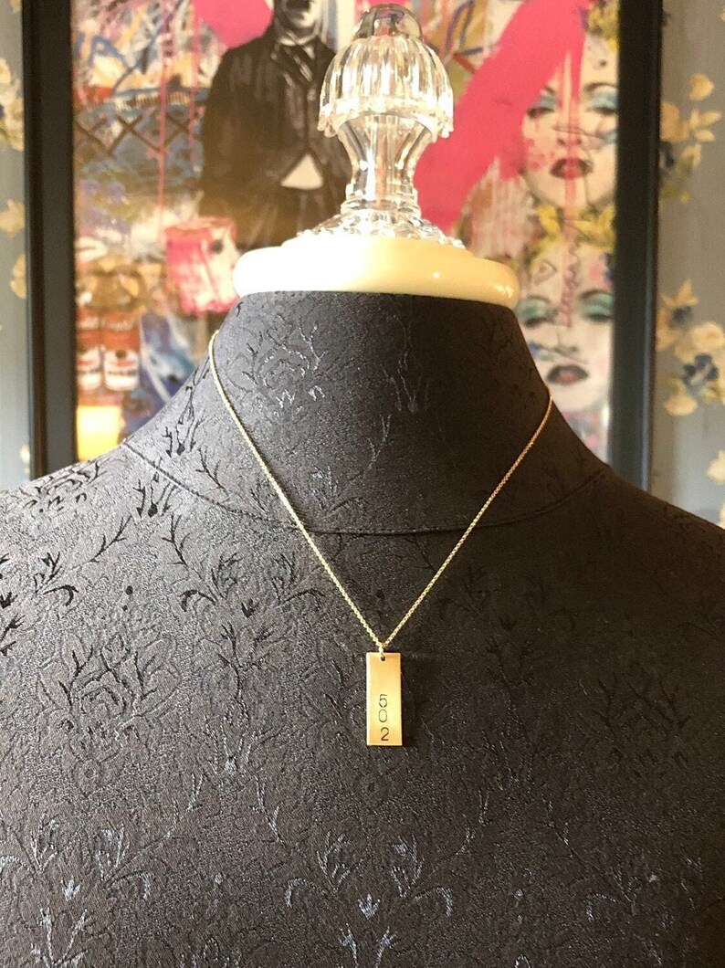 May include: A gold-colored necklace with a rectangular pendant. The pendant has the numbers "502" engraved vertically. The necklace is displayed on a black mannequin. The background includes a colorful painting.