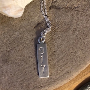 May include: A silver-colored pendant necklace with a rectangular tag. The tag has the numbers "917" etched on it. The necklace chain is delicate and silver. The background is a neutral, textured surface.