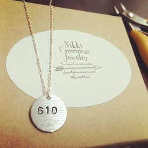 Może przedstawiać: Srebrny naszyjnik z okrągłą zawieszką z wygrawerowanym numerem "610". Zawieszka wisi na delikatnym łańcuszku. Naszyjnik jest prezentowany na brązowym pudełku z białą owalną naklejką z tekstem. Ż&oacute;łte szczypce znajdują się w tle.