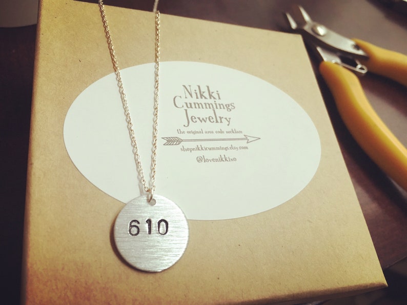 Może przedstawiać: Srebrny naszyjnik z okrągłą zawieszką z wygrawerowanym numerem "610". Zawieszka wisi na delikatnym łańcuszku. Naszyjnik jest prezentowany na brązowym pudełku z białą owalną naklejką z tekstem. Ż&oacute;łte szczypce znajdują się w tle.