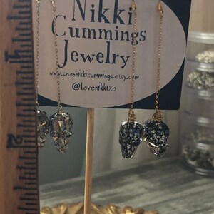 May include: Gold-toned chain earrings with skull-shaped pendants. The pendants are encrusted with sparkling crystals and dark blue and silver flecks. The earrings hang from gold-colored ear wires. The sign behind the earrings reads "Nikki Cummings Jewelry".