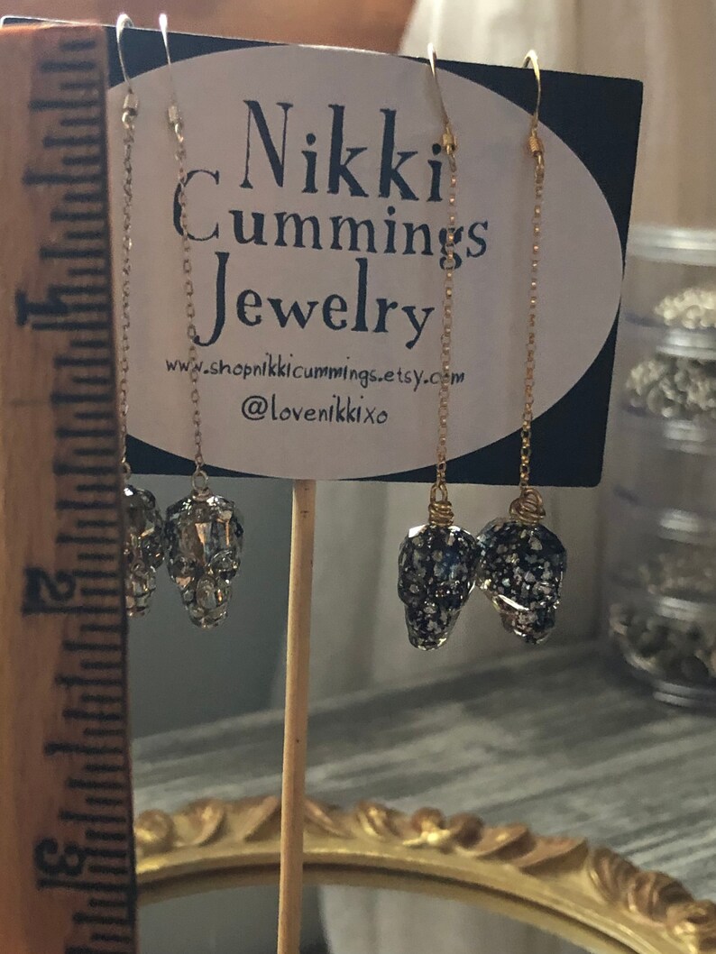 May include: Gold-toned chain earrings with skull-shaped pendants. The pendants are encrusted with sparkling crystals and dark blue and silver flecks. The earrings hang from gold-colored ear wires. The sign behind the earrings reads "Nikki Cummings Jewelry".