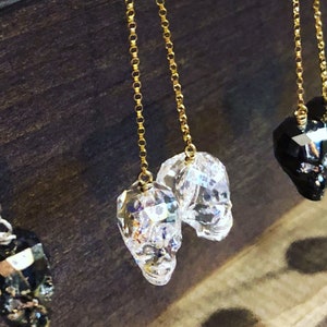 May include: Clear crystal skull earrings with gold-colored chain. The earrings are faceted, catching the light and creating a sparkling effect. The skulls are detailed and have a gothic aesthetic.