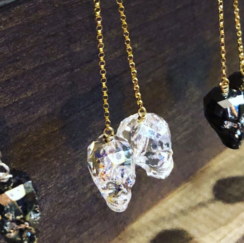May include: Clear crystal skull earrings with gold-colored chain. The earrings are faceted, catching the light and creating a sparkling effect. The skulls are detailed and have a gothic aesthetic.
