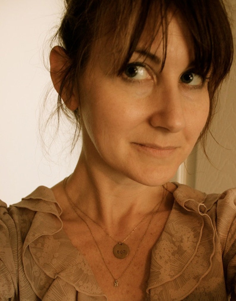 Może przedstawiać: Kobieta z brązowymi włosami i oczami, ubrana w beżową bluzkę z falbankowym dekoltem. Ma na sobie dwa złote naszyjniki, jeden z okrągłą zawieszką z numerem "502" i drugi z małym urokiem.