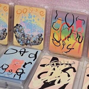 Puede incluir: Un conjunto de seis fundidos de cera en un embalaje de plástico transparente. Cada fundido presenta un diseño abstracto diferente con fondos coloridos y líneas negras. Los diseños incluyen "pogue", "dad" y "love you lots".