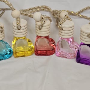 May include: Five heart-shaped glass bottles with wooden tops and string hangers. The bottles are filled with colored liquid and are arranged in a row. The colors are blue, yellow, pink, light pink, and purple.