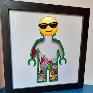 May include: Framed artwork featuring a yellow emoji with sunglasses above a green Lego figure outline. The Lego figure's body is filled with colorful Lego pieces and accessories. The frame is black.