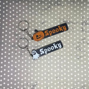 May include: Two black and white keychains with the word "Spooky" written on them. One keychain has a pumpkin charm and the other has a ghost charm.