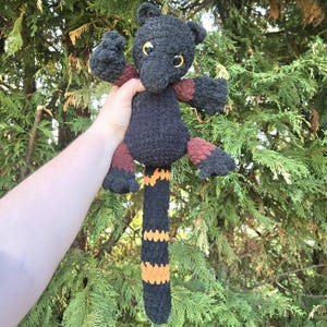 Peut inclure: Un animal en crochet noir fait à la main avec une longue queue rayée et des yeux jaunes. Les bras et les jambes sont accentués de marron et de noir. La peluche est tenue devant un fond de feuillage vert.