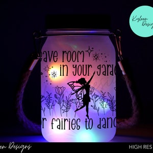 May include: A white glass jar with a black silhouette of a fairy and flowers. The jar has a rope handle and lights up with multi-coloured LEDs. The text on the jar reads "Leave room in your garden for fairies to dance".